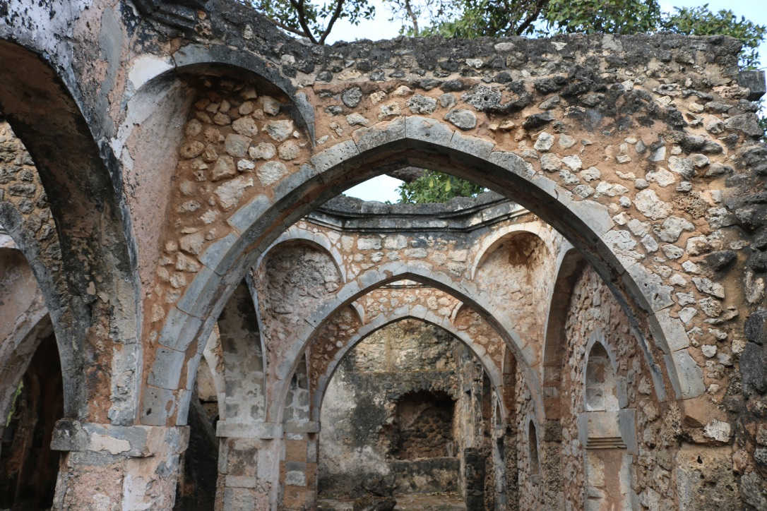 Sunlight enters the ruined coral-stone mosque interior through pointed arches and roof openings at Kilwa Kisiwani.