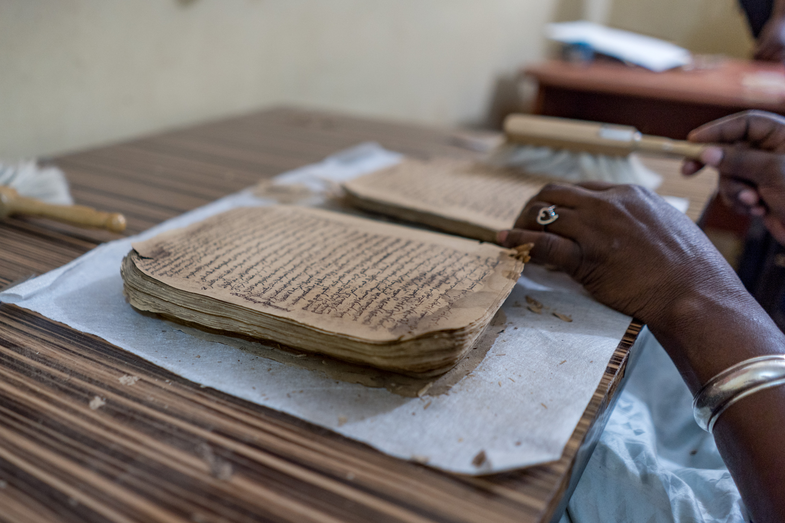 White-gloved hands use a soft brush to clean an Arabic manuscript page on a conservation work surface.