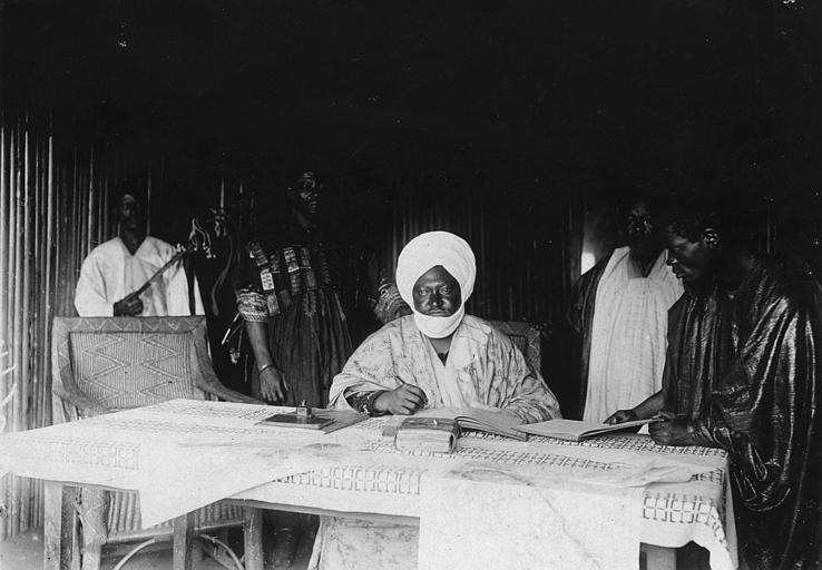 Ibrahim Njoya sits with a pen at a table holding an open book, surrounded by several attendants.